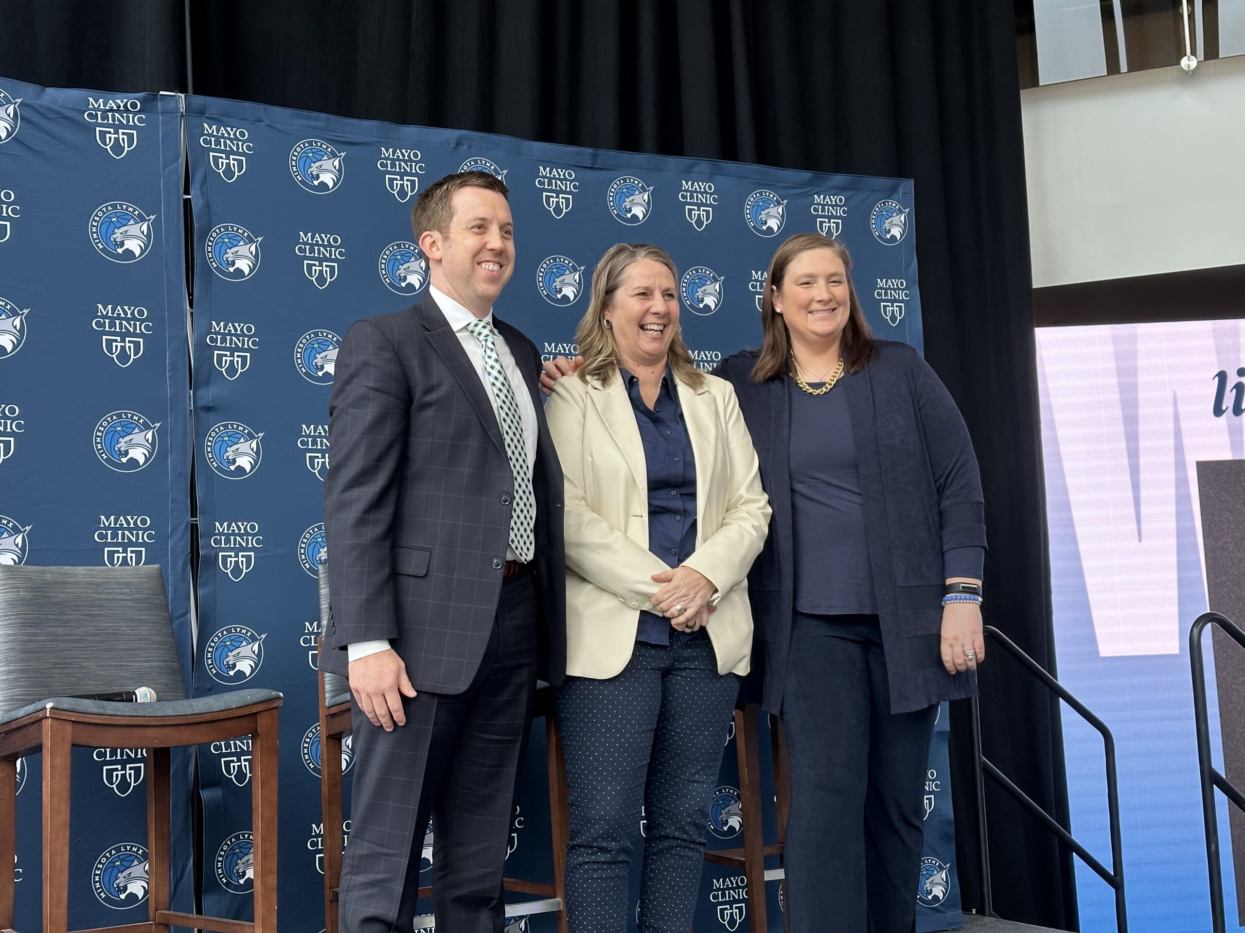 Minnesota Lynx coaches Eric Thibault, Cheryl Reeve and Lindsay Whalen pose for a photo while standing on a podium in front of a blue backdrop with Lynx logos.