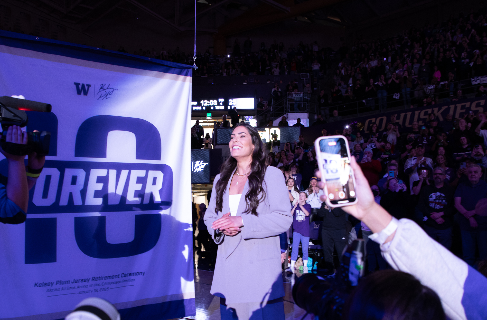 Kelsey Plum looks up to the rafters as the University of Washington retires her No. 10 to the rafters in Alaska Airlines Arena. She stands in front of a 'Forever 10' banner as all sorts of media crowd to take photos.