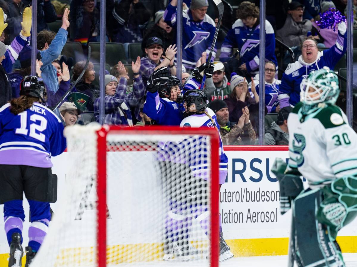 Minnesota players celebrate with a hug behind the Boston goal. They are wearing purple home uniforms.