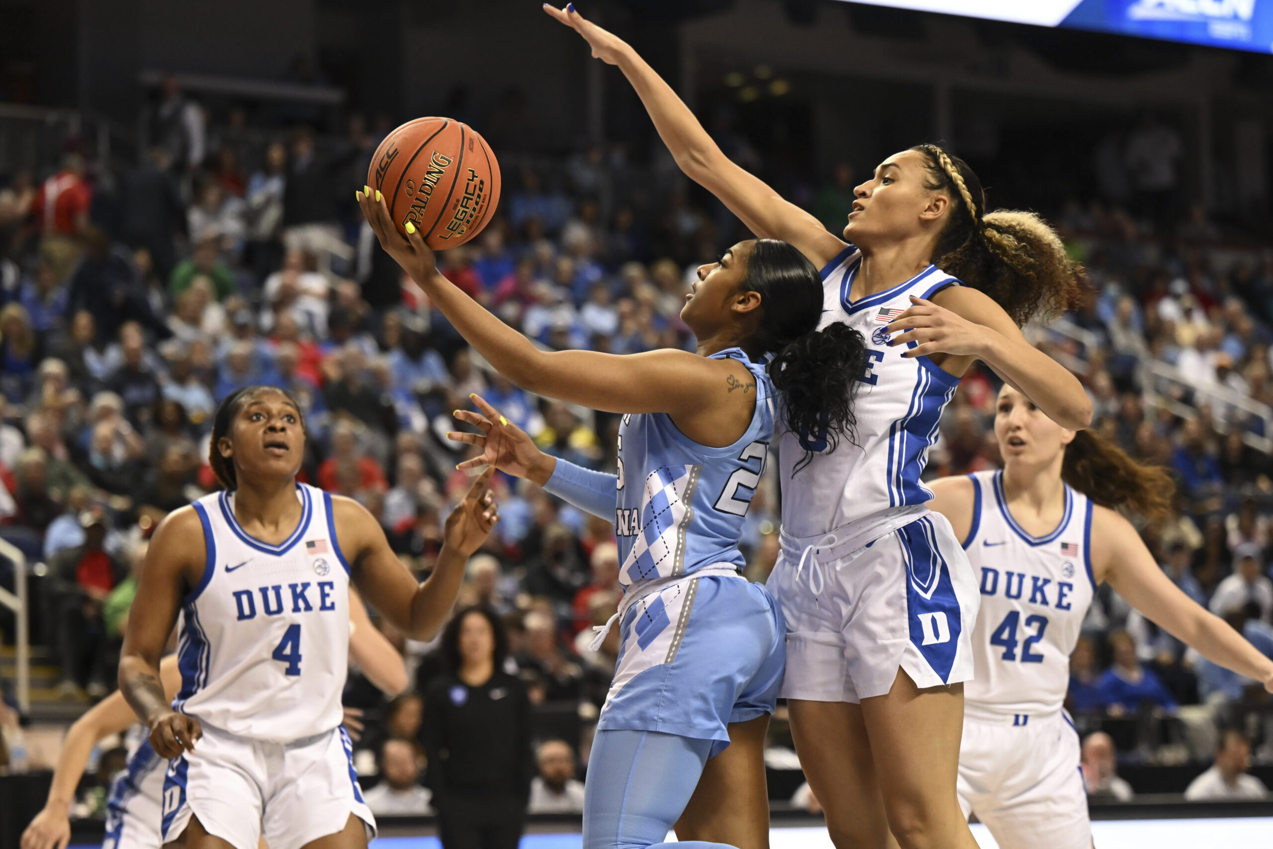 North Carolina guard Deja Kelly raises the ball in her hand to attempt a scoop layup while Duke guard Celeste Taylor jumps from her side to block the shot. They are playing in an installation of the Tobacco road rivalry with teammates in the midground and a packed crowd in the background