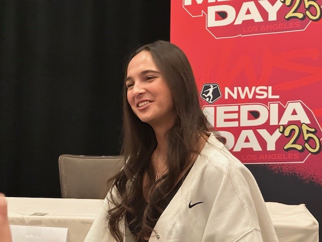 A woman smiles in front of an NWSL media day banner