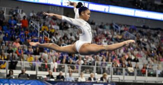 A gymnast in a white leotard executes a split leap on the balance beam.