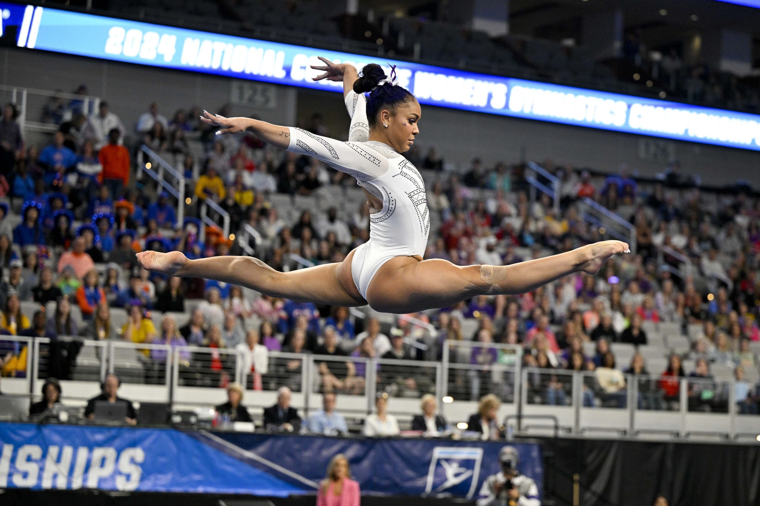 A gymnast in a white leotard executes a split leap on the balance beam.