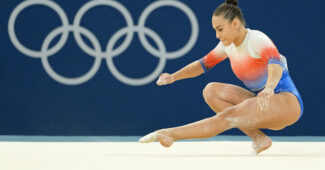 A gymnast in a red, white, and blue leotard performs a floor exercise in front of the Olympic rings.
