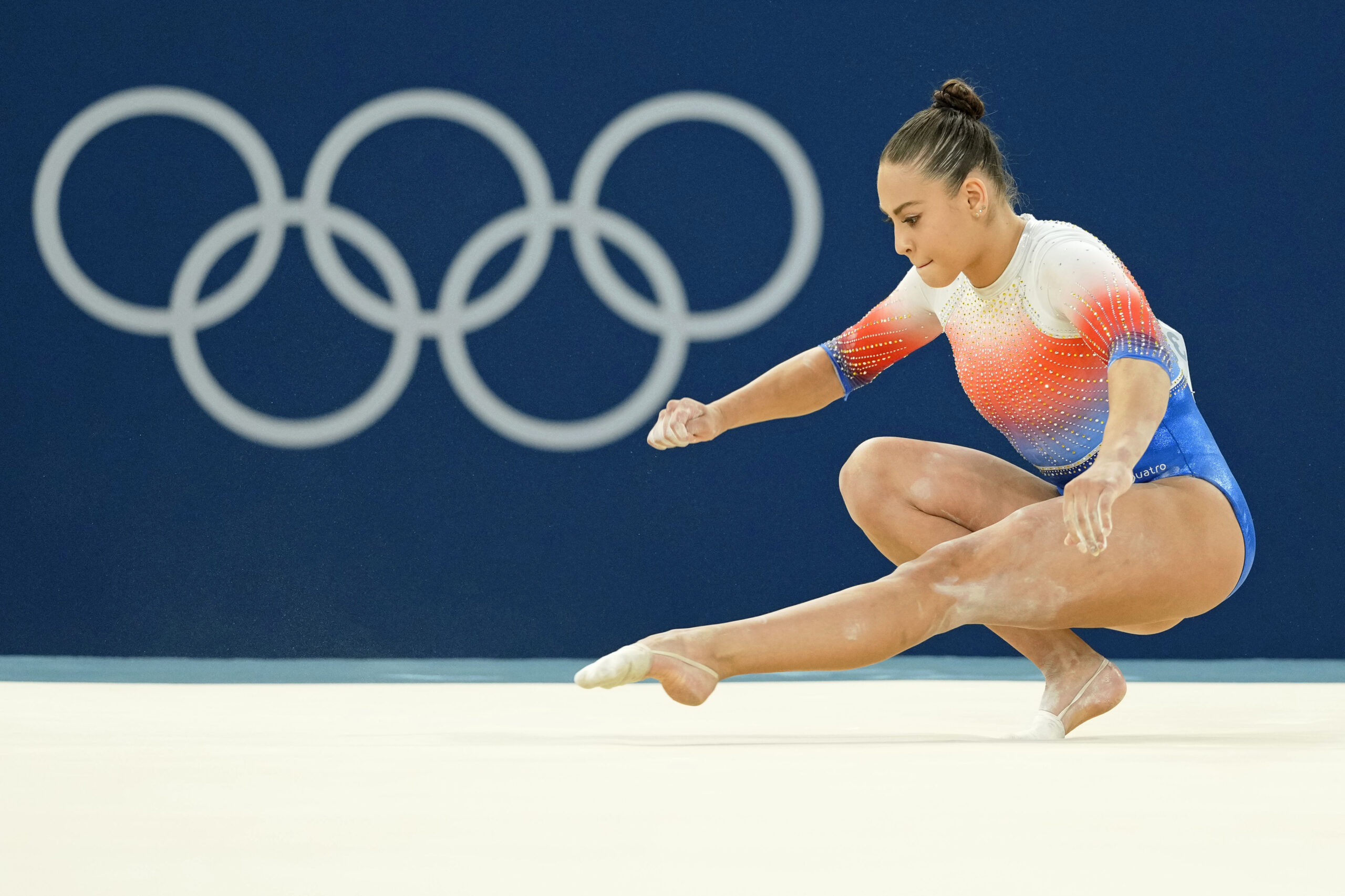 A gymnast in a red, white, and blue leotard performs a floor exercise in front of the Olympic rings.