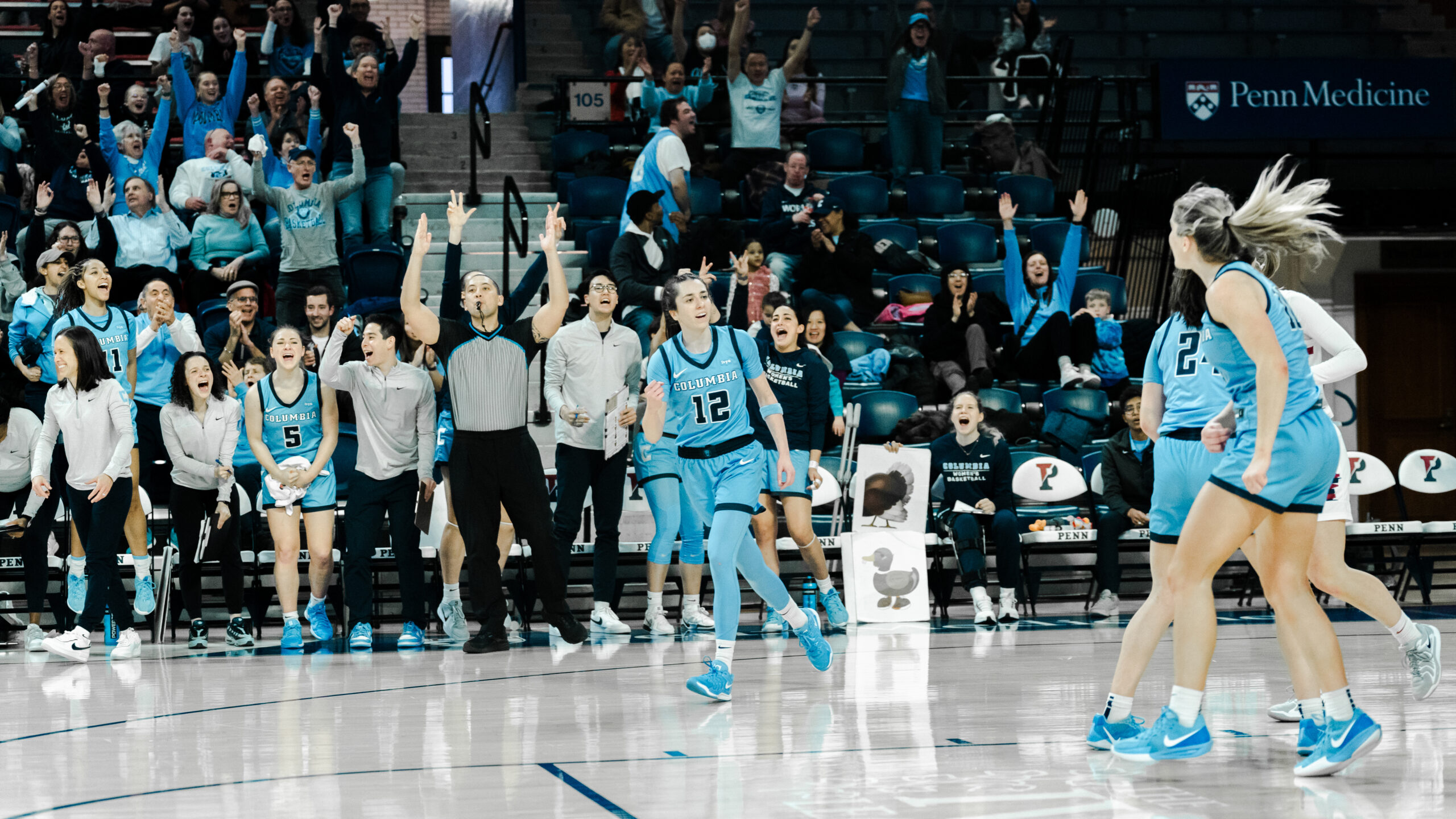 Columbia guard Marija Avlijas pumps her right fist as the Lions' bench and fans behind her celebrate. The referee, also behind her, has both hands up to signal a 3-pointer.
