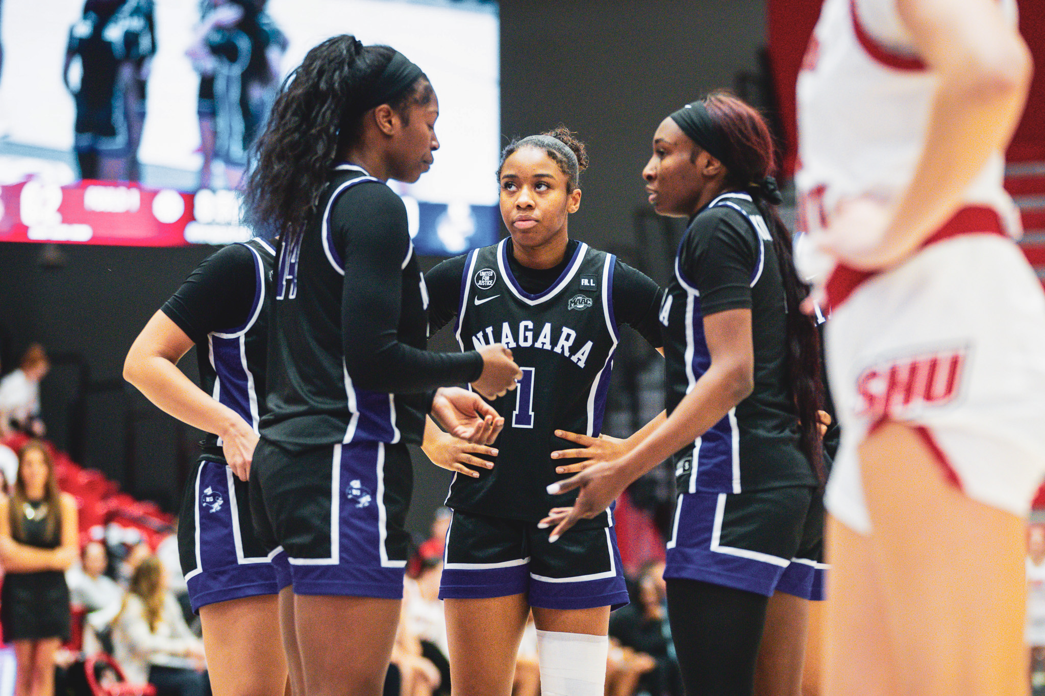 Four Niagara women's basketball players (in black) huddle around each other while freshman guard Nevaeh Otis (No. 1, center) looks to her right and has her hands on her hips. (Photo credit: Demetrius Kazanas).