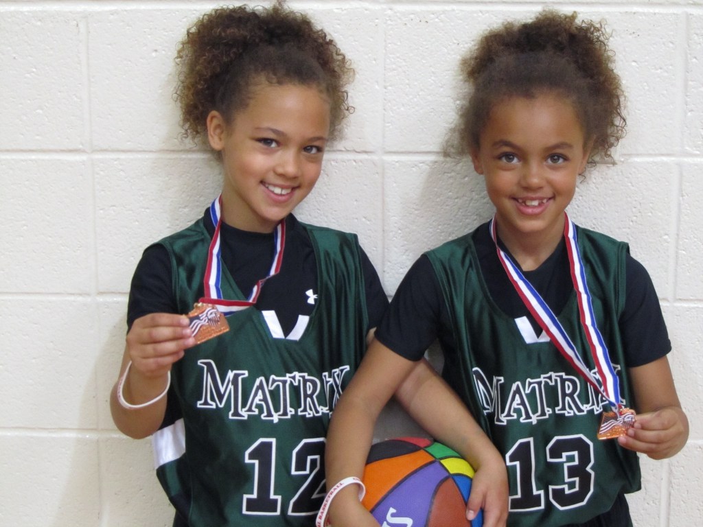 Grace and Alayna Arnolie smile for a photo in dark green jerseys with the team name "Matrix" on them. Medals with an American flag theme are around each of their necks. Together, they're holding a rainbow basketball.