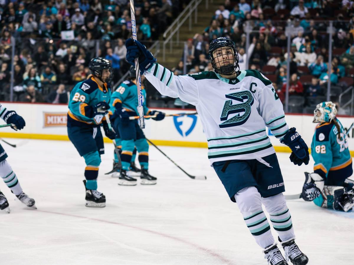 Knight raises her stick in celebration. She is facing the camera and wearing a white away uniform.