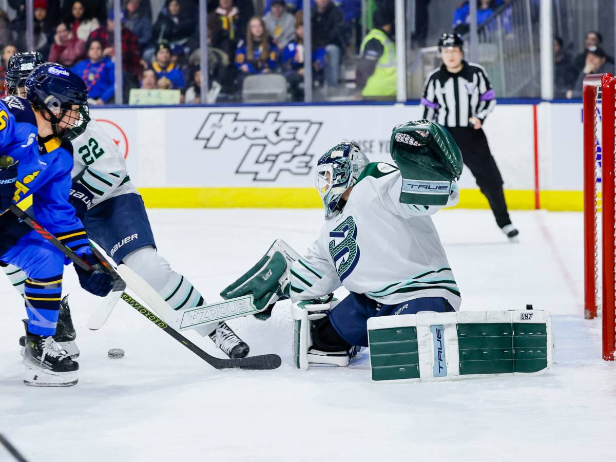 From her knees, Frankel pushes to the side and kicks the puck out to her right. She is wearing a white away uniform.