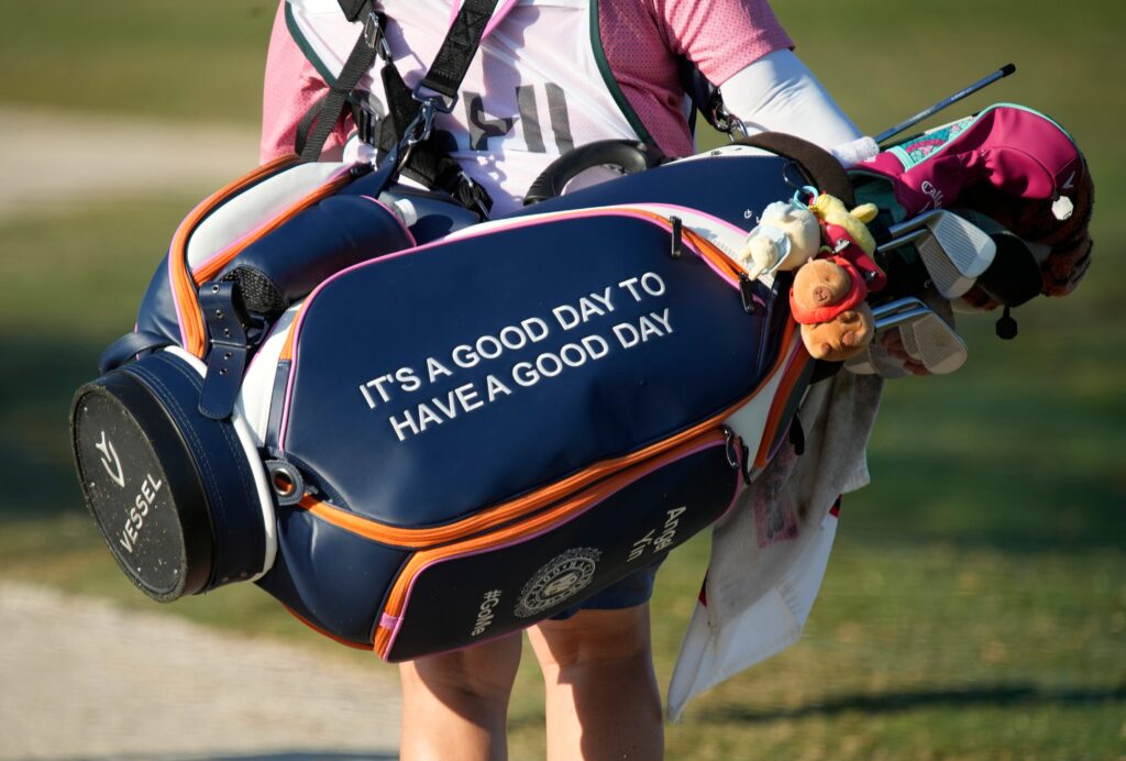 A golf caddie carrying a golf bag with the phrase "It's a good day to have a good day" walks forward with their hands supporting the bag.