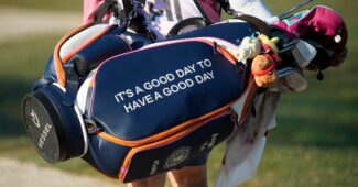 A golf caddie carrying a golf bag with the phrase "It's a good day to have a good day" walks forward with their hands supporting the bag.