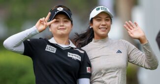 Two golfers smile and acknowledge the camera, one on left holds up two fingers in a peace sign and one on right waves with left hand.