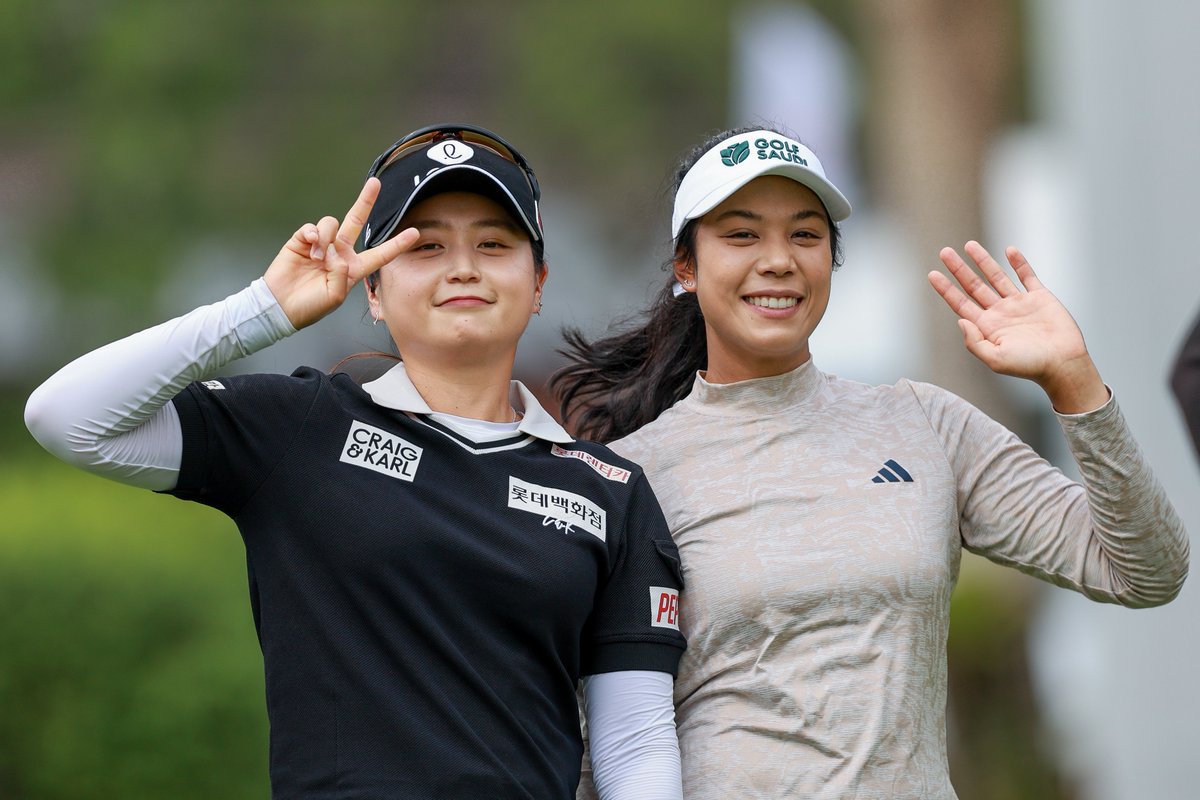 Two golfers smile and acknowledge the camera, one on left holds up two fingers in a peace sign and one on right waves with left hand.