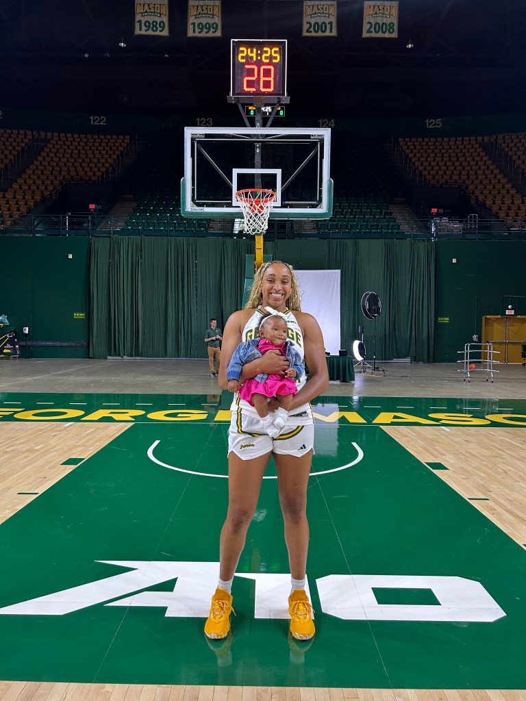 George Mason forward Nalani Kaysia, dressed in her white George Mason uniform, smiles while holding her smiling daughter Xéla on the Atlantic 10 logo on the court of EagleBank Arena.