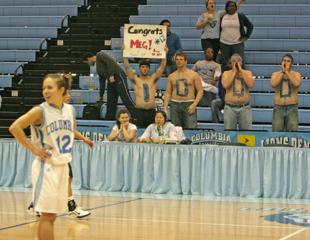 Columbia guard Megan Griffith stands on the court and looks off to the side. In the background, four men have painted their chests to spell out "1,000," and one of them has a sign reading, "Congrats Meg! #12."