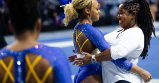 A coach in a white shirt is hugging a gymnast in a blue leotard with yellow straps. Another gymnast is in the foreground facing them.