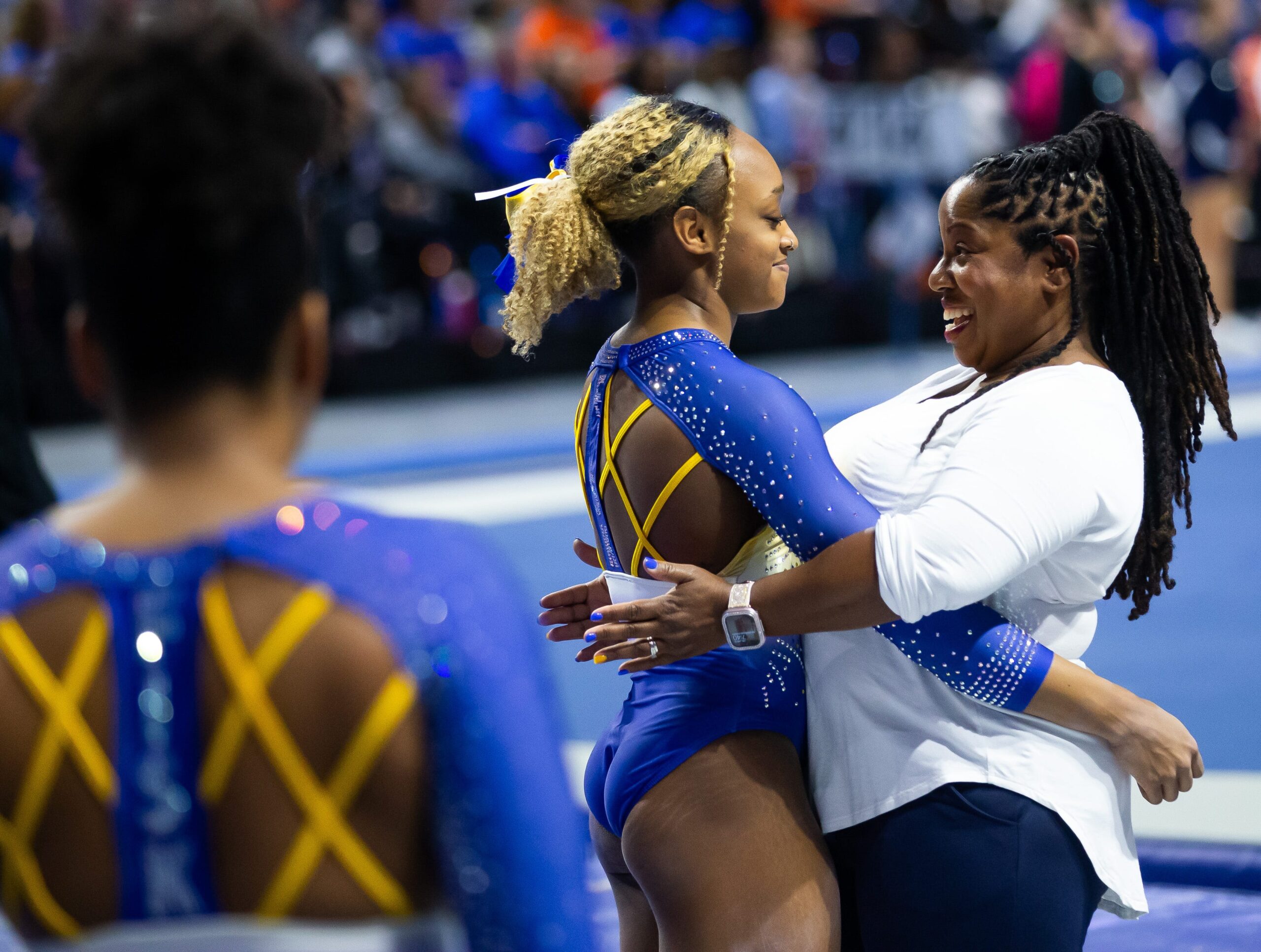 A coach in a white shirt is hugging a gymnast in a blue leotard with yellow straps. Another gymnast is in the foreground facing them.