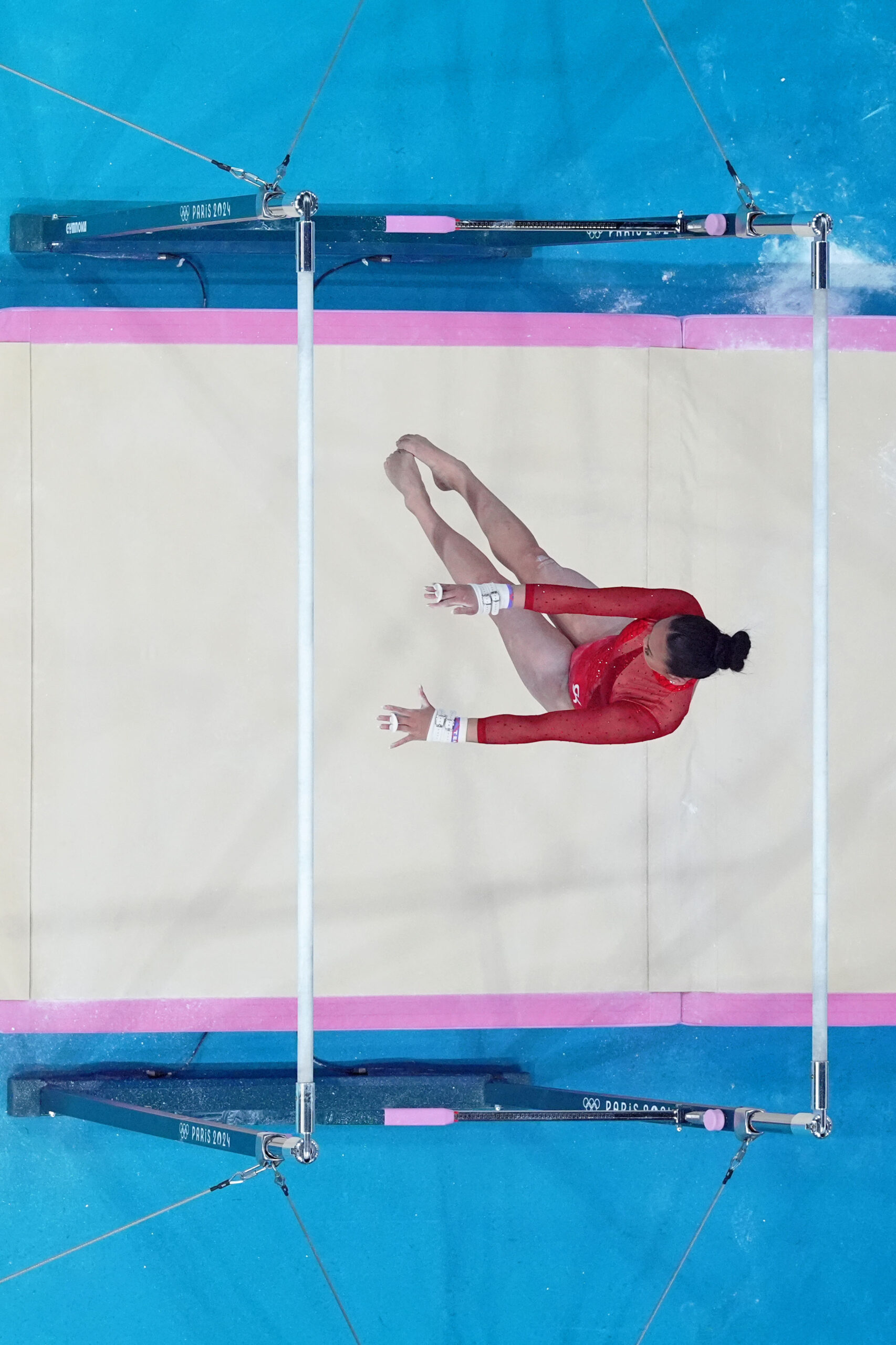 A gymnast in a red leotard competing on the uneven bars over a white mat.