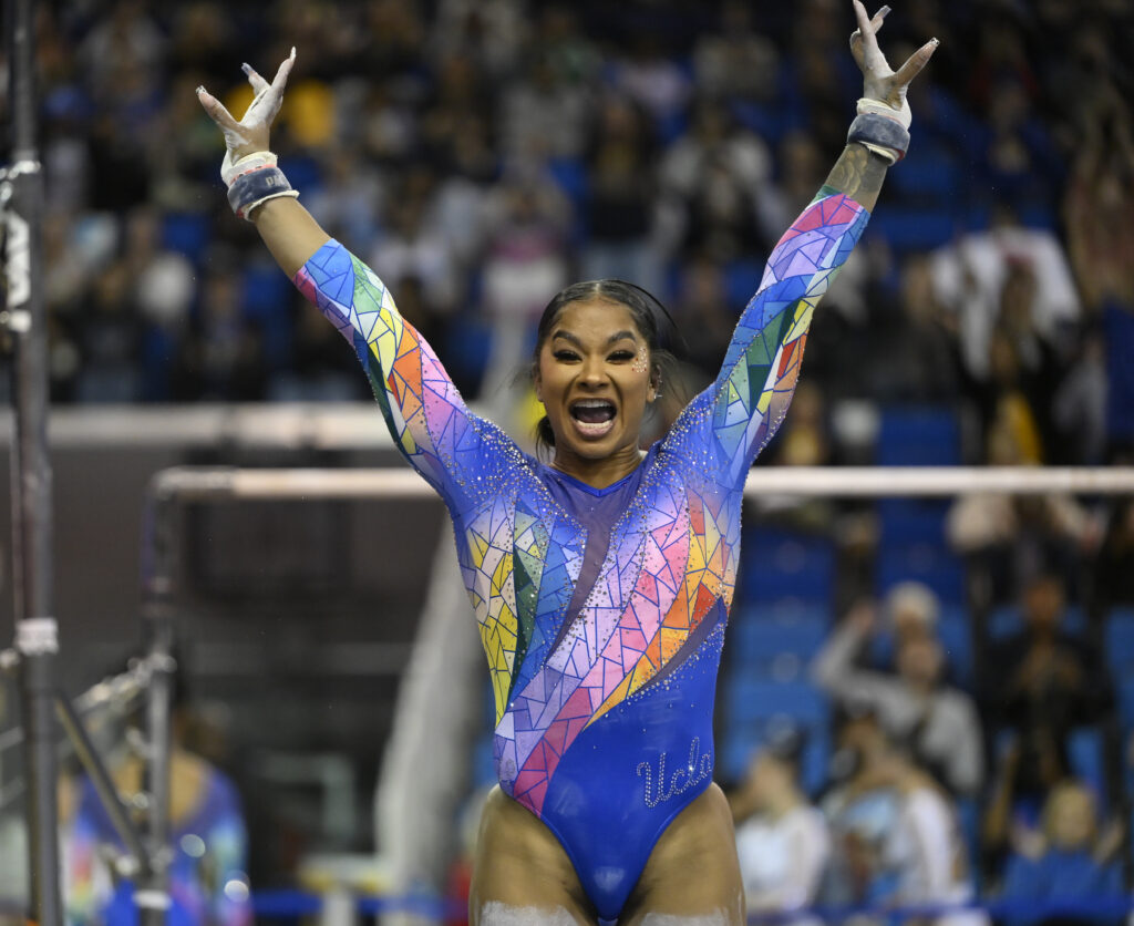 UCLA gymnast Jordan Chiles, wearing a rainbow leotard, smiles widely and salutes the judges after an uneven bars routine.
