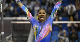 UCLA gymnast Jordan Chiles, wearing a rainbow leotard, smiles widely and salutes the judges after an uneven bars routine.