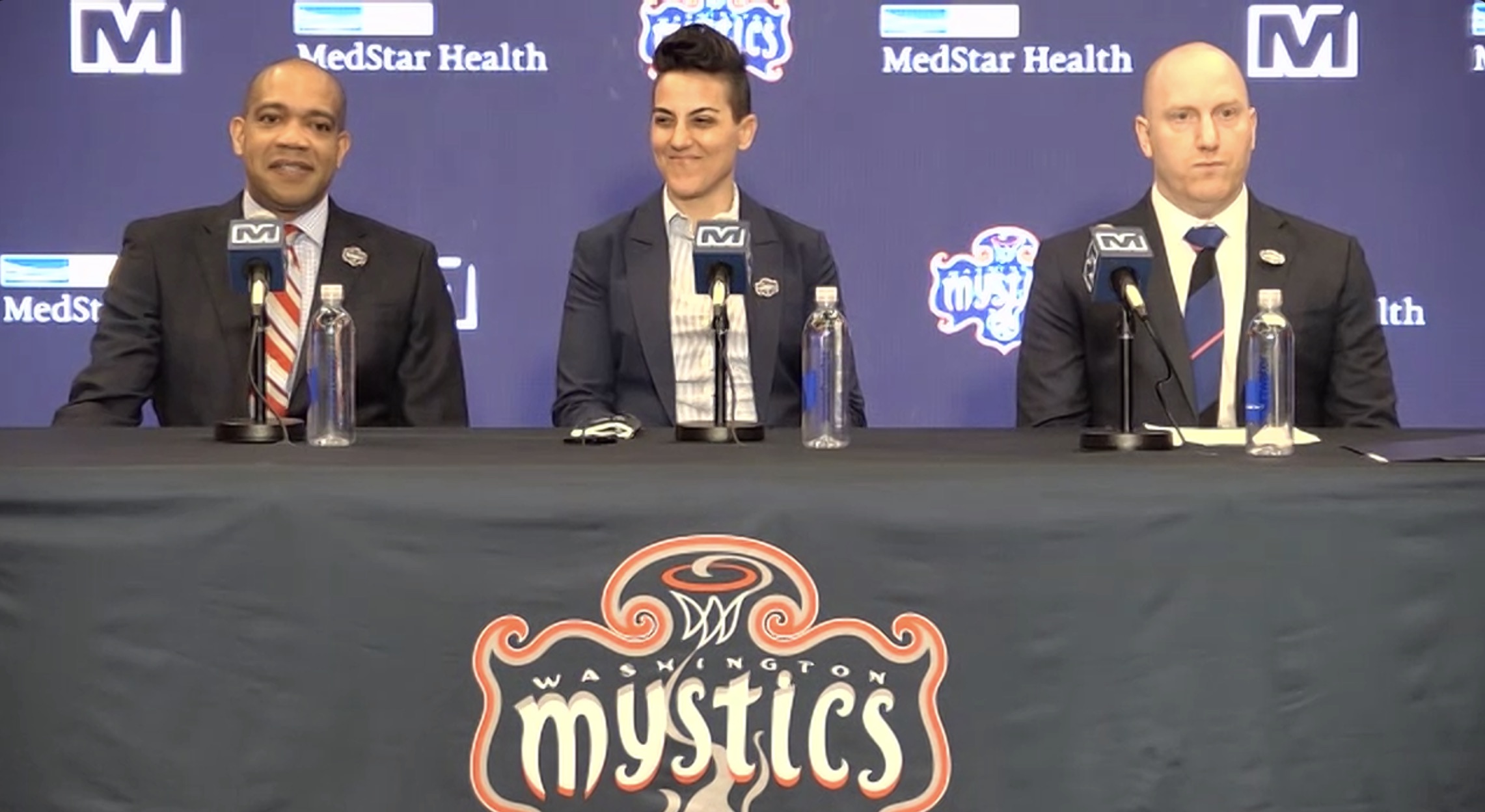 Washington Mystics head coach Sydney Johnson, Mystics general manager Jamila Wideman and Monumental Basketball president Michael Winger sit at a podium for a press conference. All three are in dark suit jackets with Mystics pins on the collars. Johnson and Wideman are smiling, while Winger stares straight ahead.