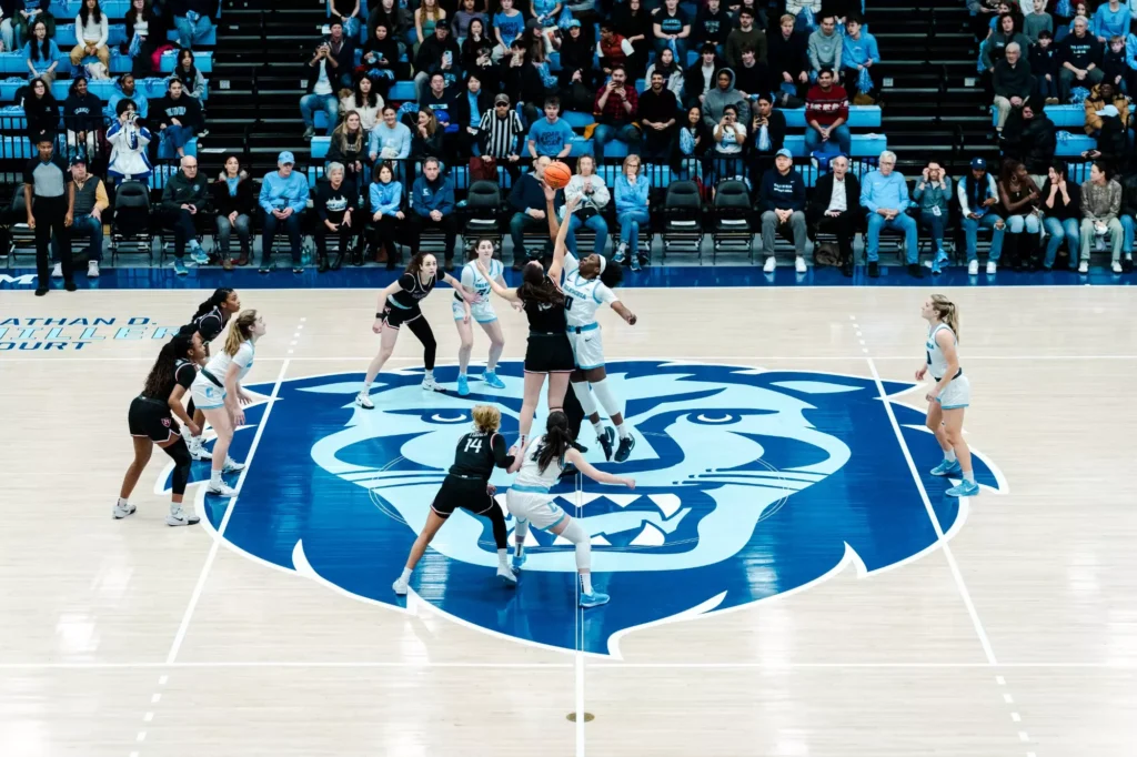One player each from Columbia and Harvard jumps in the air to contest the opening tip. The other players stand around the circle at center court, watching the ball's path.
