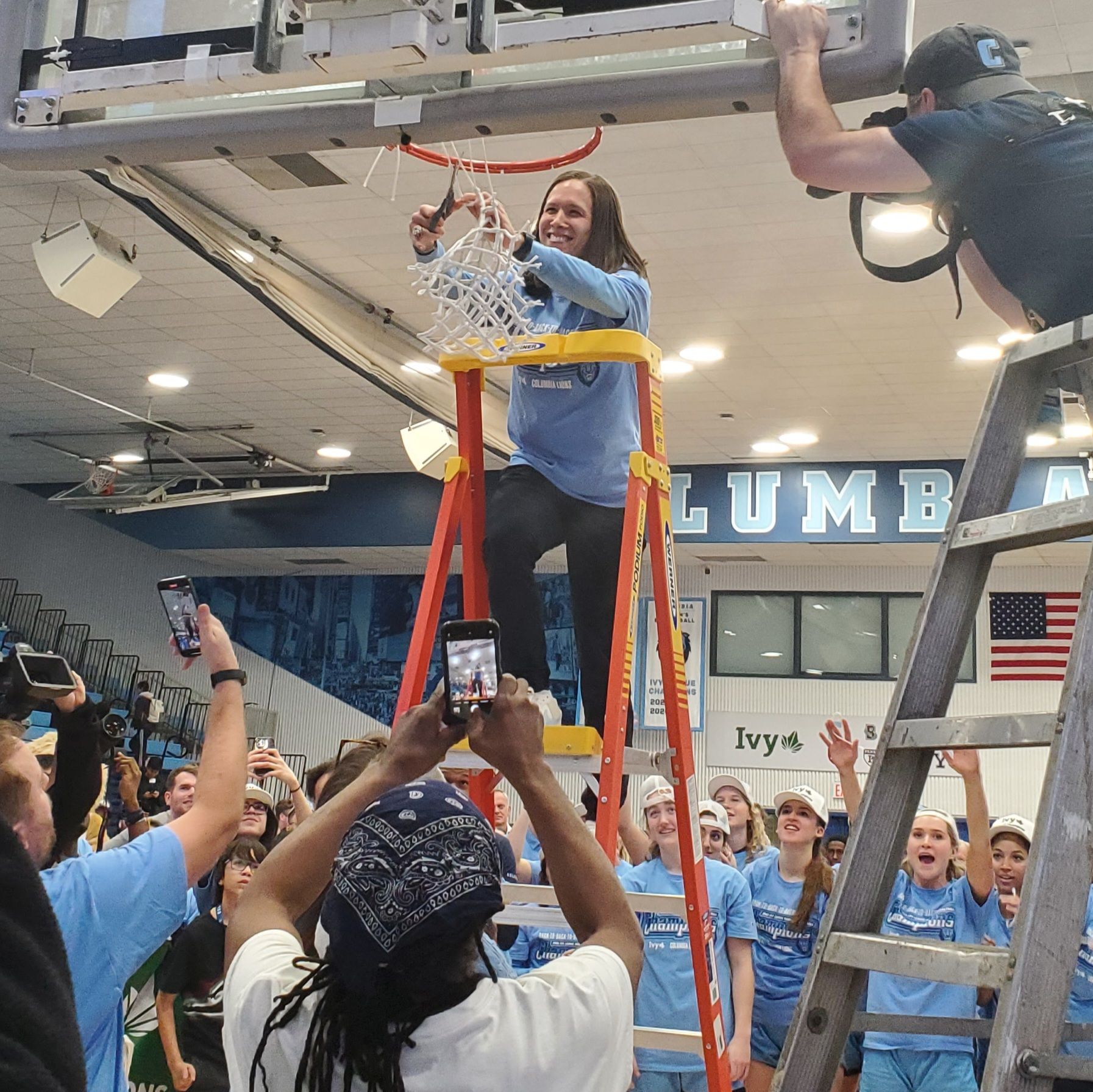 Columbia women's basketball head coach Megan Griffith stands on a ladder and cuts the net of a basketball hoop at the Columbia Lions home court while Lions players stand behind her celebrating and spectators stand in front of her taking pictures with their phones