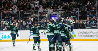 Fleet players celebrate with a tight group hug while the crowd cheers in the background. They are wearing green home uniforms.