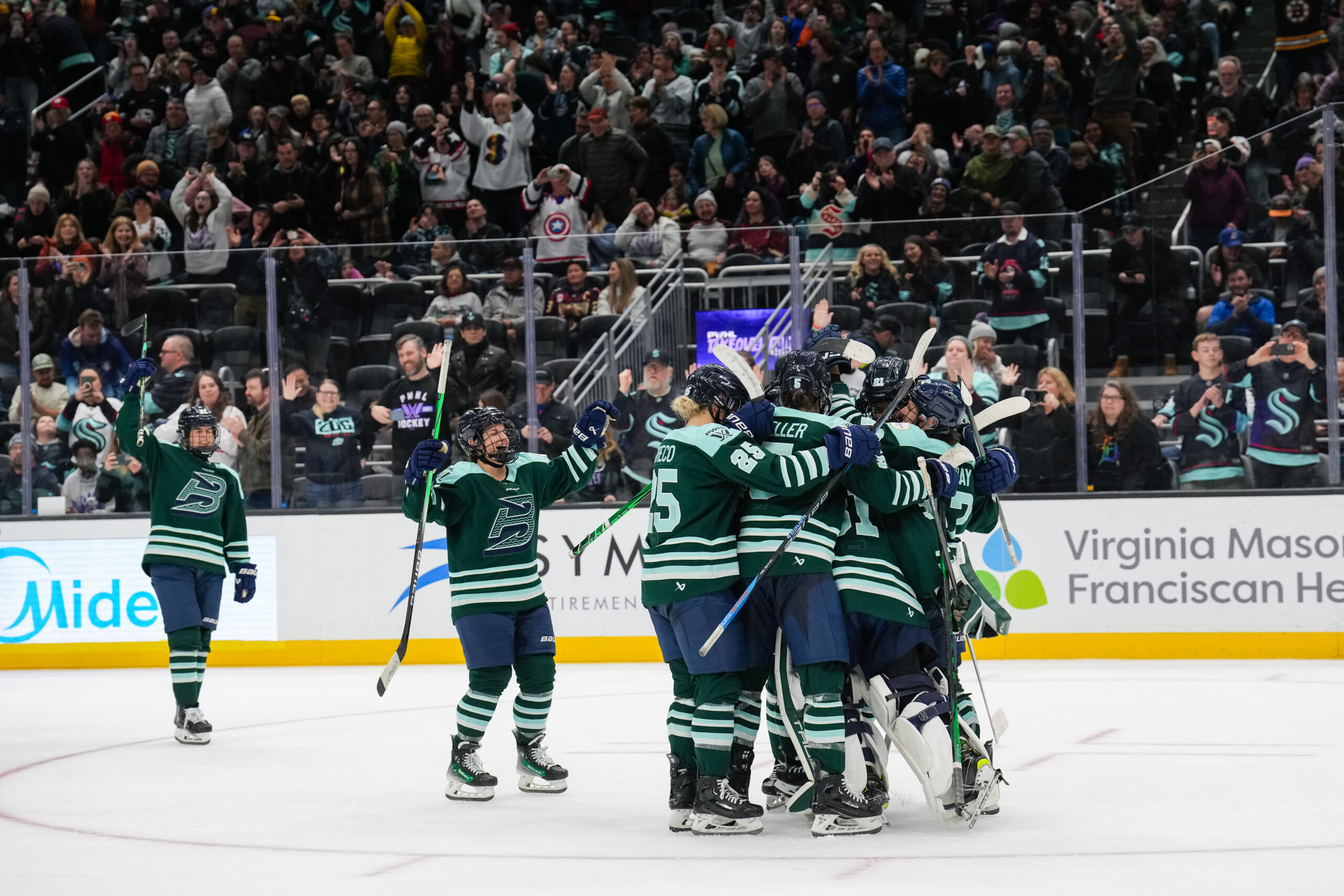 Fleet players celebrate with a tight group hug while the crowd cheers in the background. They are wearing green home uniforms.