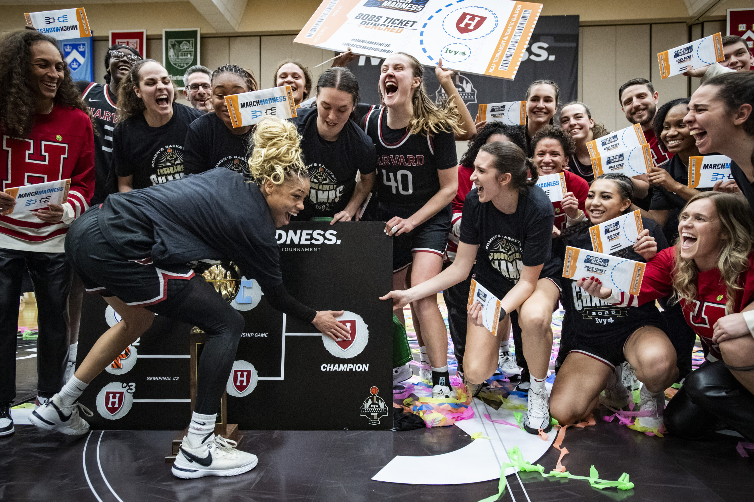 Harvard guard Harmoni Turner puts a Harvard sticker on the Ivy League Tournament bracket after winning the event. Behind her, her teammates cheer and hold up mini "tickets" celebrating their qualification for the NCAA Tournament.