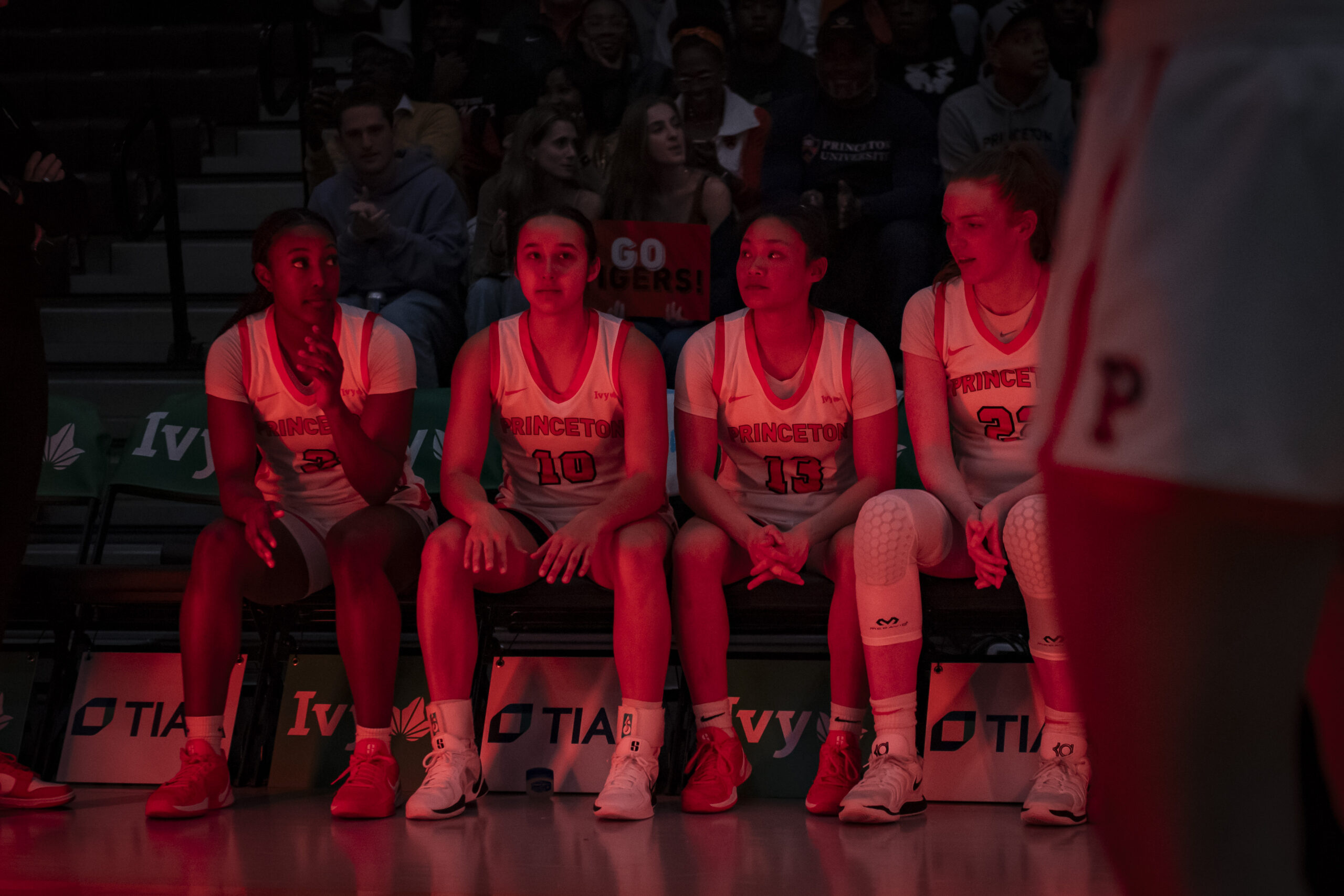 Princeton guard/forward Olivia Hutcherson, guard Skye Belker, guard Ashley Chea and forward Parker Hill sit on the Princeton bench and wait to hear their names called as starters. The arena is dark and there is a reddish glow from the lighting and special effects.