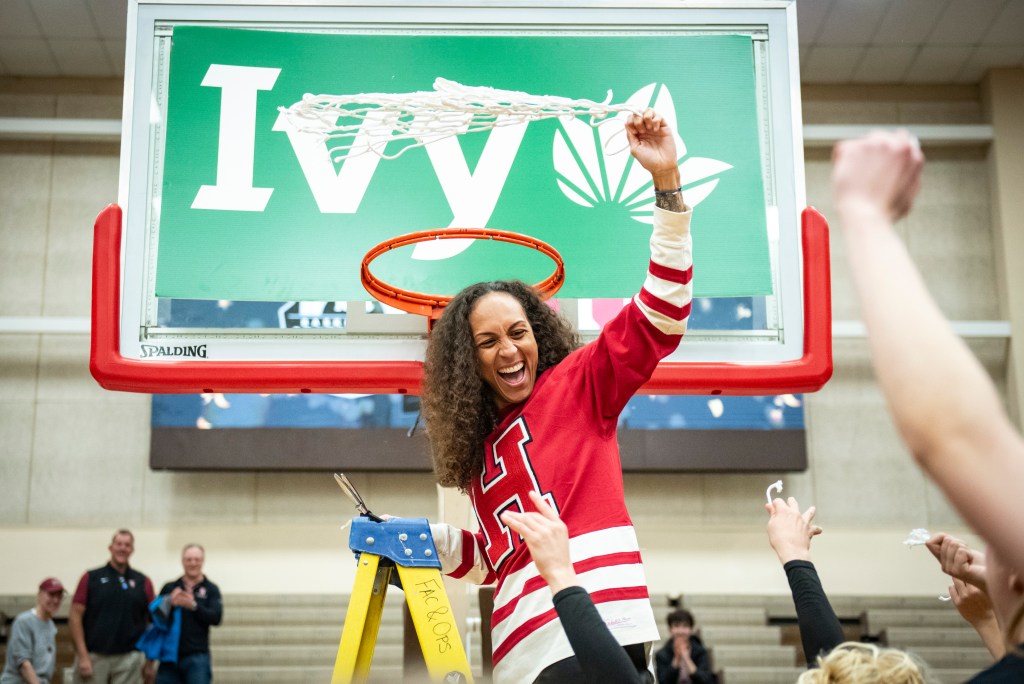 Harvard head coach Carrie Moore stands on a ladder holding scissors in her right hand. In her left hand, she twirls the net she just cut down above her head. She looks at her players, who are cheering from the ground.