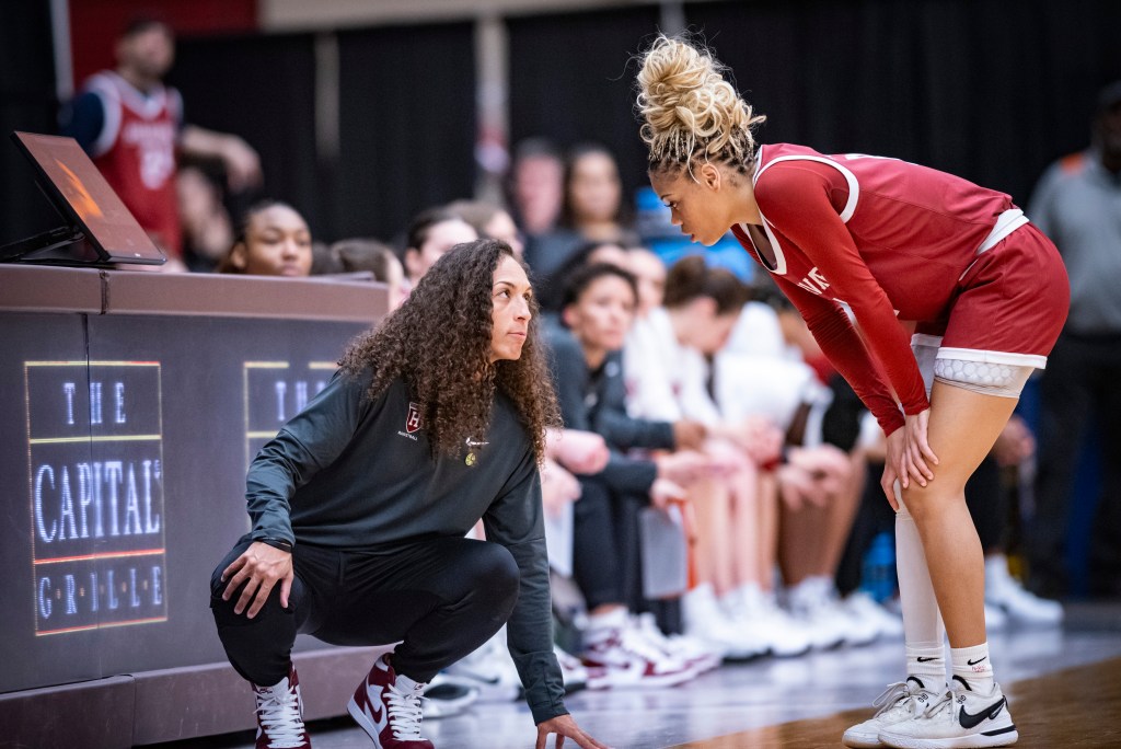 Harvard head coach Carrie Moore squats on the sideline and looks up at guard Harmoni Turner. Turner stands in bounds during a stoppage in play. She leans over to talk to Moore with her hands on her knees.