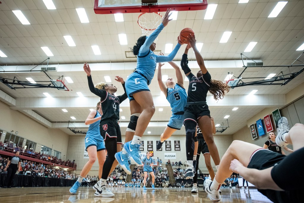 Columbia guard/forward Perri Page uses her left hand to deflect a layup attempt by Harvard guard Gabby Anderson on the left side of the rim. Columbia guard Mia Broom is also leaping in the air behind Anderson to help contest the shot.
