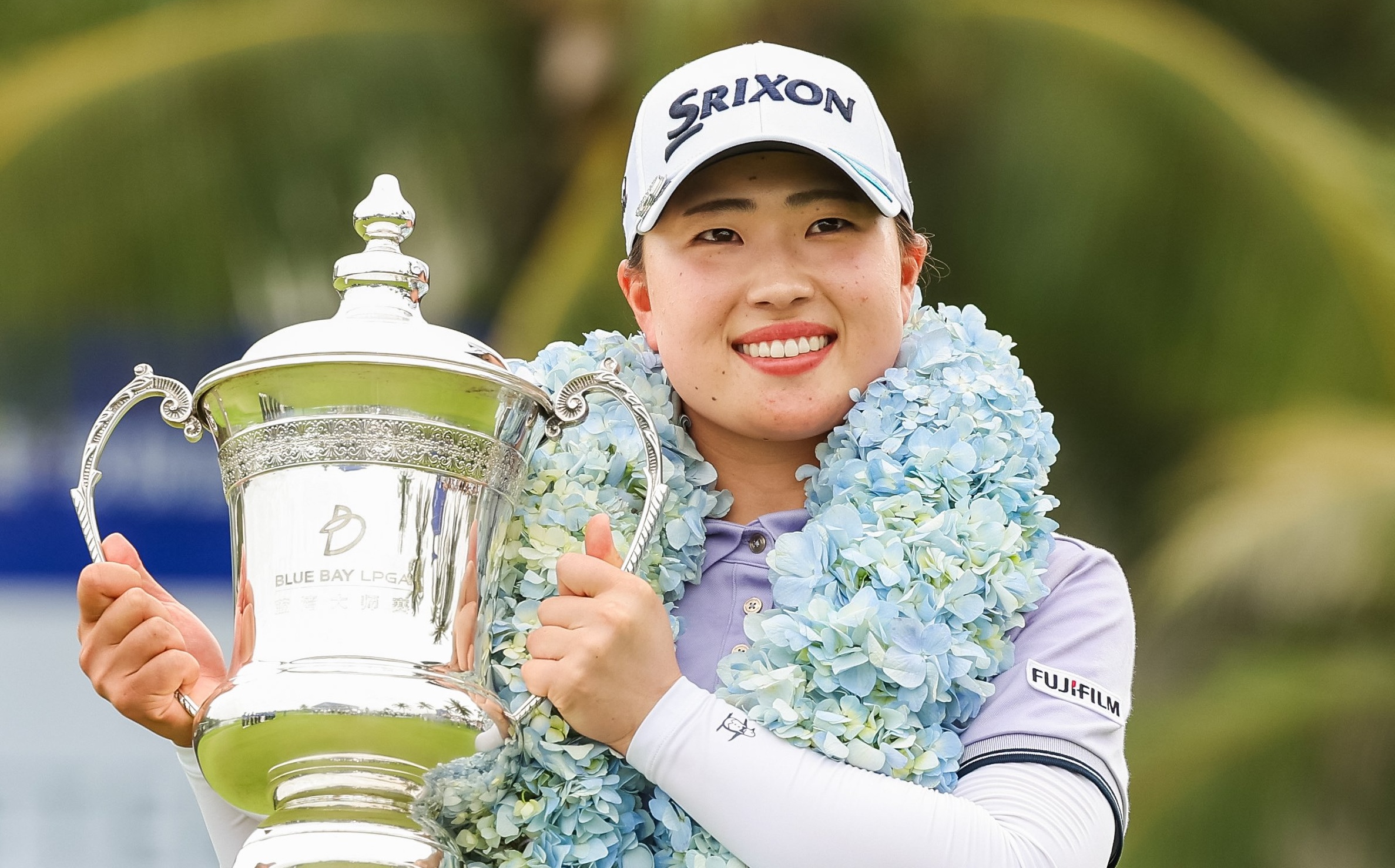 Rio Takeda, wearing a flowered boa, holds the Blue Bay LPGA trophy to her right side and smiles.