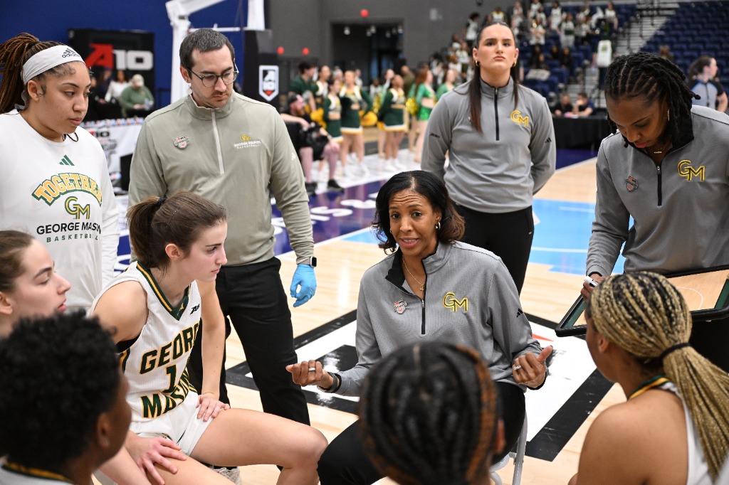 A George Mason assistant draws up a play while her head coach sits and talks to the players who are in a semicircle in front of her.