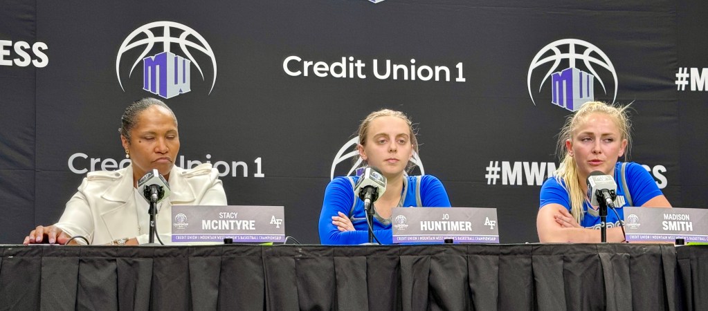 A coach and two basketball players sits behind a table and answer questions at a post-game press conference.