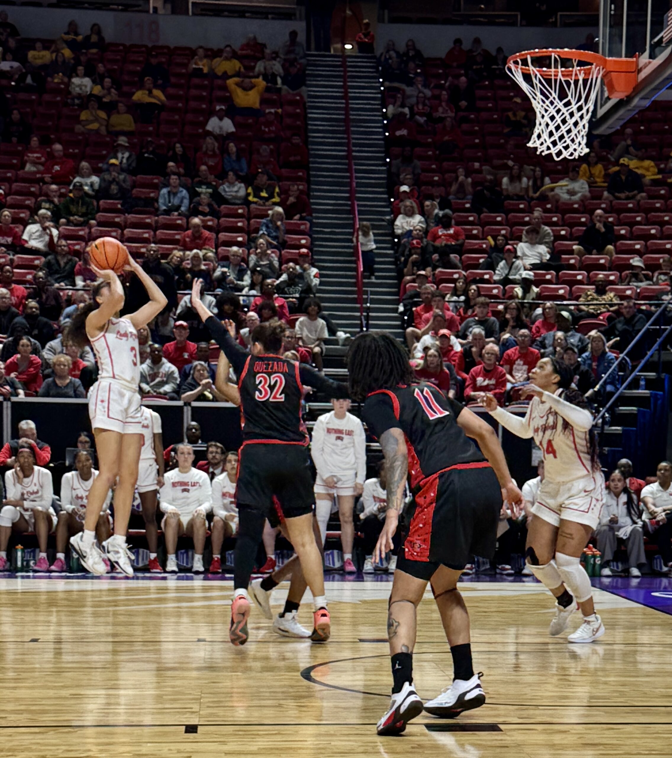 A basketball player shoots a jump shot over the out-stretched hand of a defender.