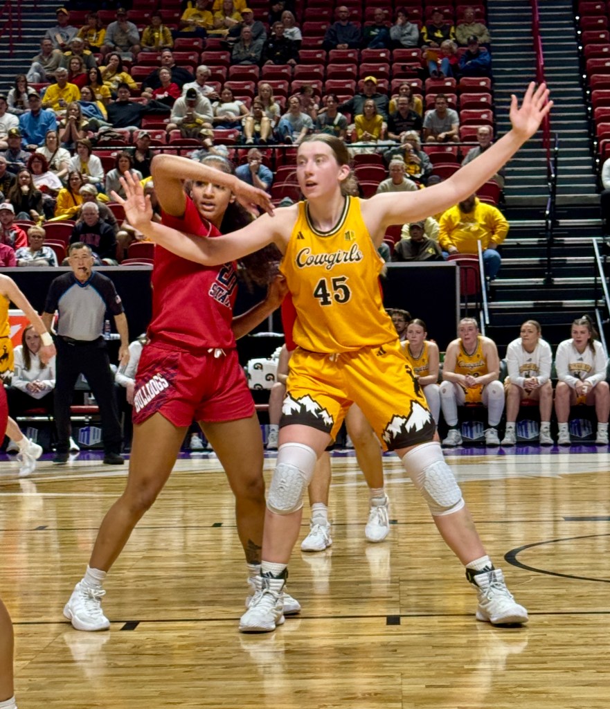 A basketball player facing the camera extends her arms in anticipation of receiving a pass from a teammate with a defender on her right hip fighting for position