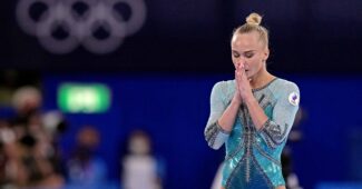 A gymnast in a green leotard pauses before a routine in front of the Olympic rings symbol.