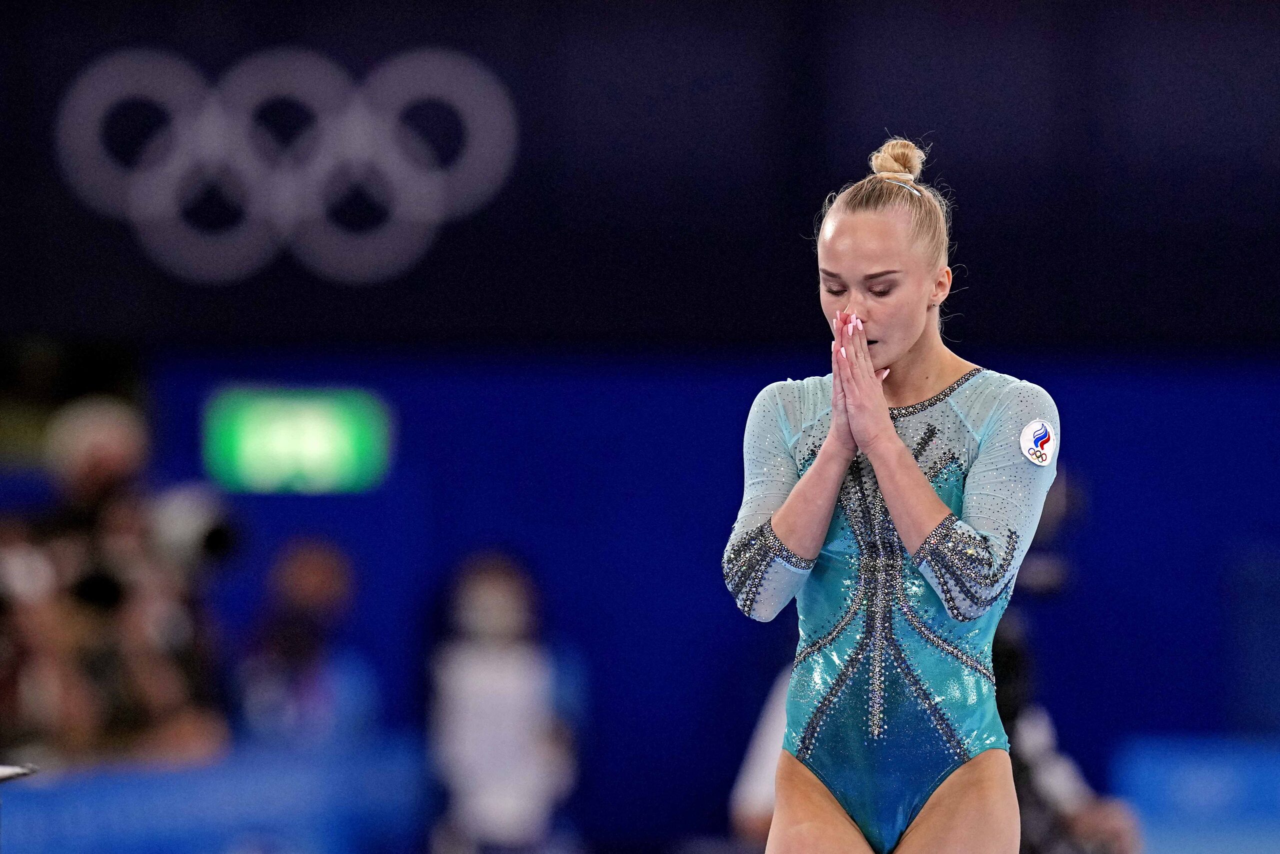 A gymnast in a green leotard pauses before a routine in front of the Olympic rings symbol.