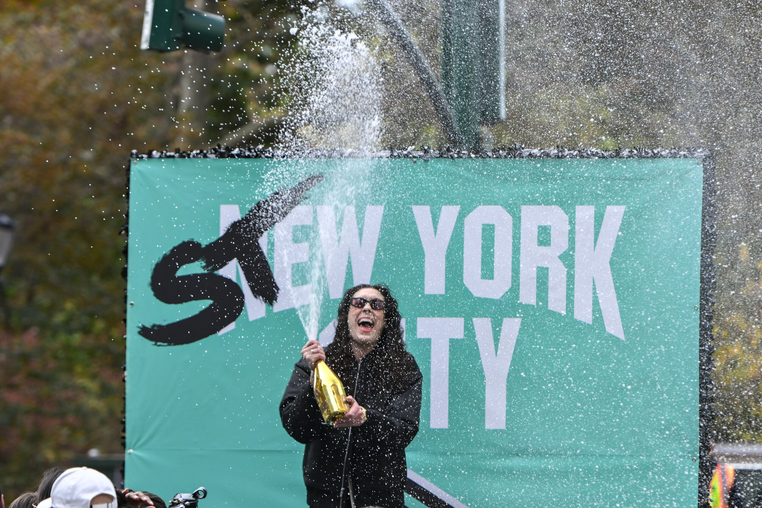 Breanna Stewart celebrates with a bottle of Champagne behind a seafoam background that says Stew York City.
