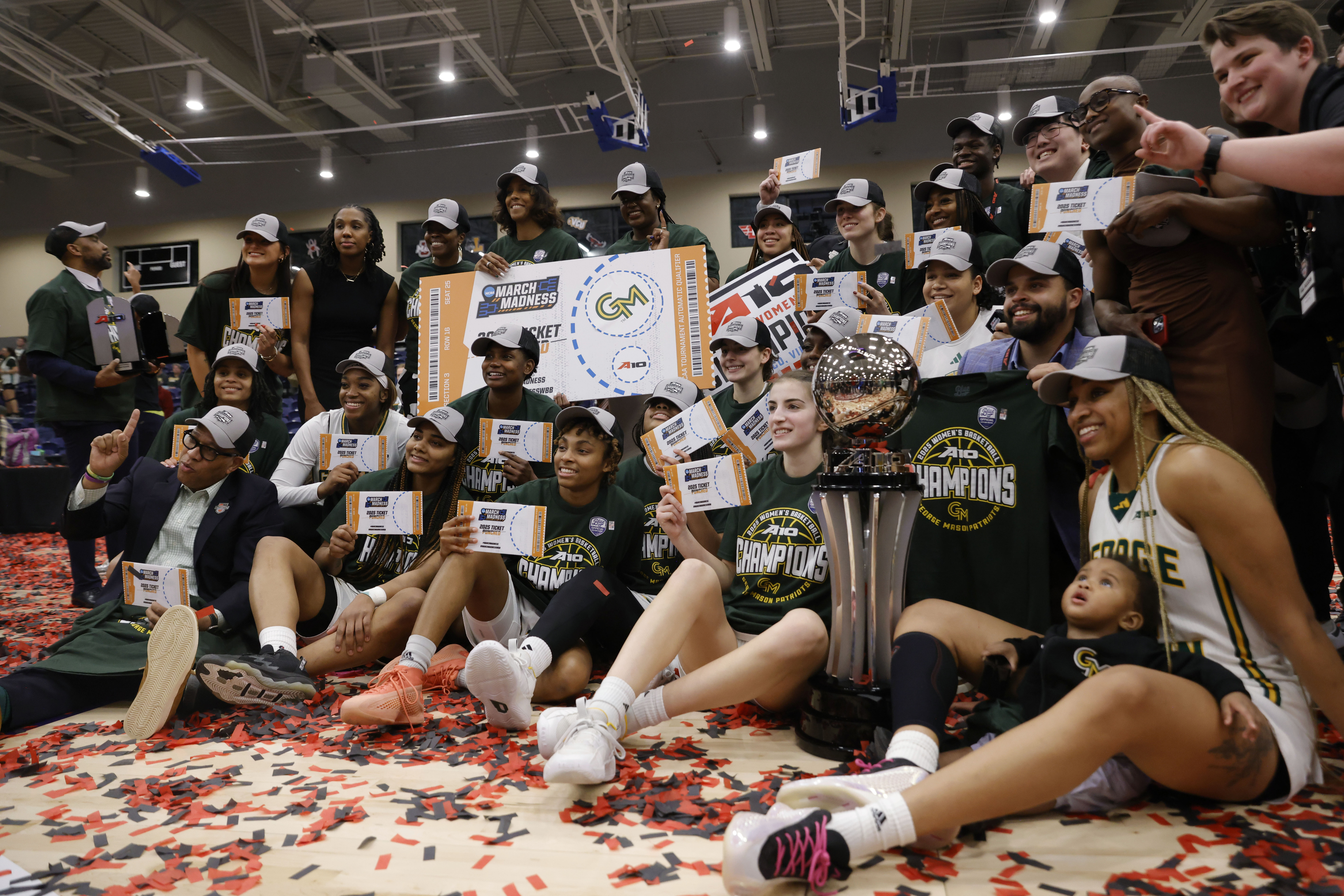 George Mason poses with the the Atlantic 10 championship trophy, the team’s oversized ticket to March Madness, smaller handheld tickets, All-Championship team trophies and and a championship logo sign. The team is wearing its championship hats and shirts or holding them up.