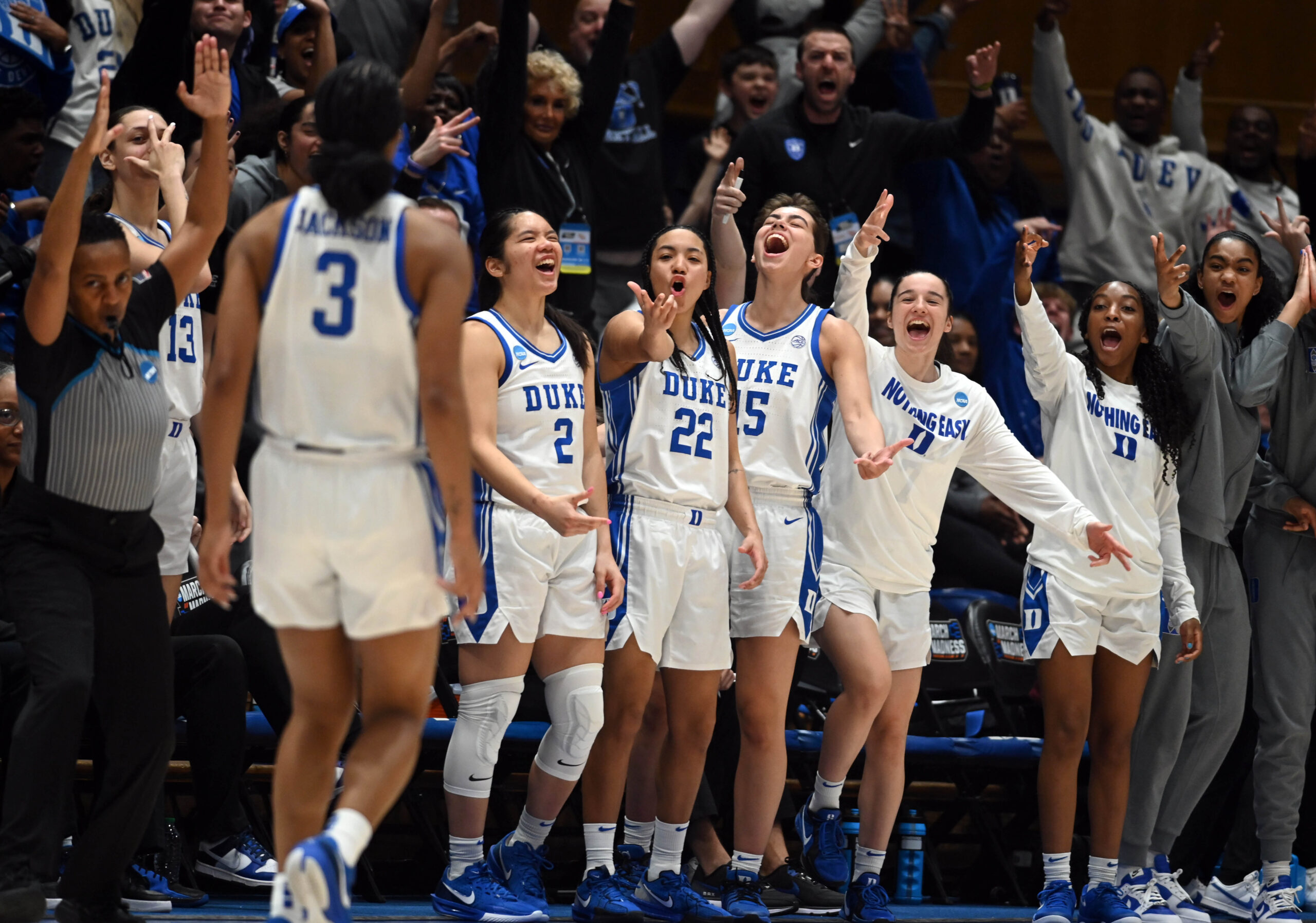 Ashlon Jackson turns toward seven players standing and celebrating at the Duke bench, including Jordan Wood holding her hand in a number-3 gesture in front of her face.