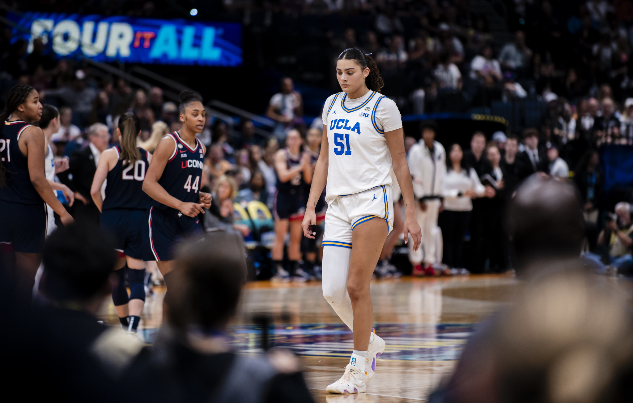 UCLA center Lauren Betts walks off the floor during a Final Four game. She looks down at the court. Fans and the Final Four's "Four It All" slogan are visible but out of focus in the background.