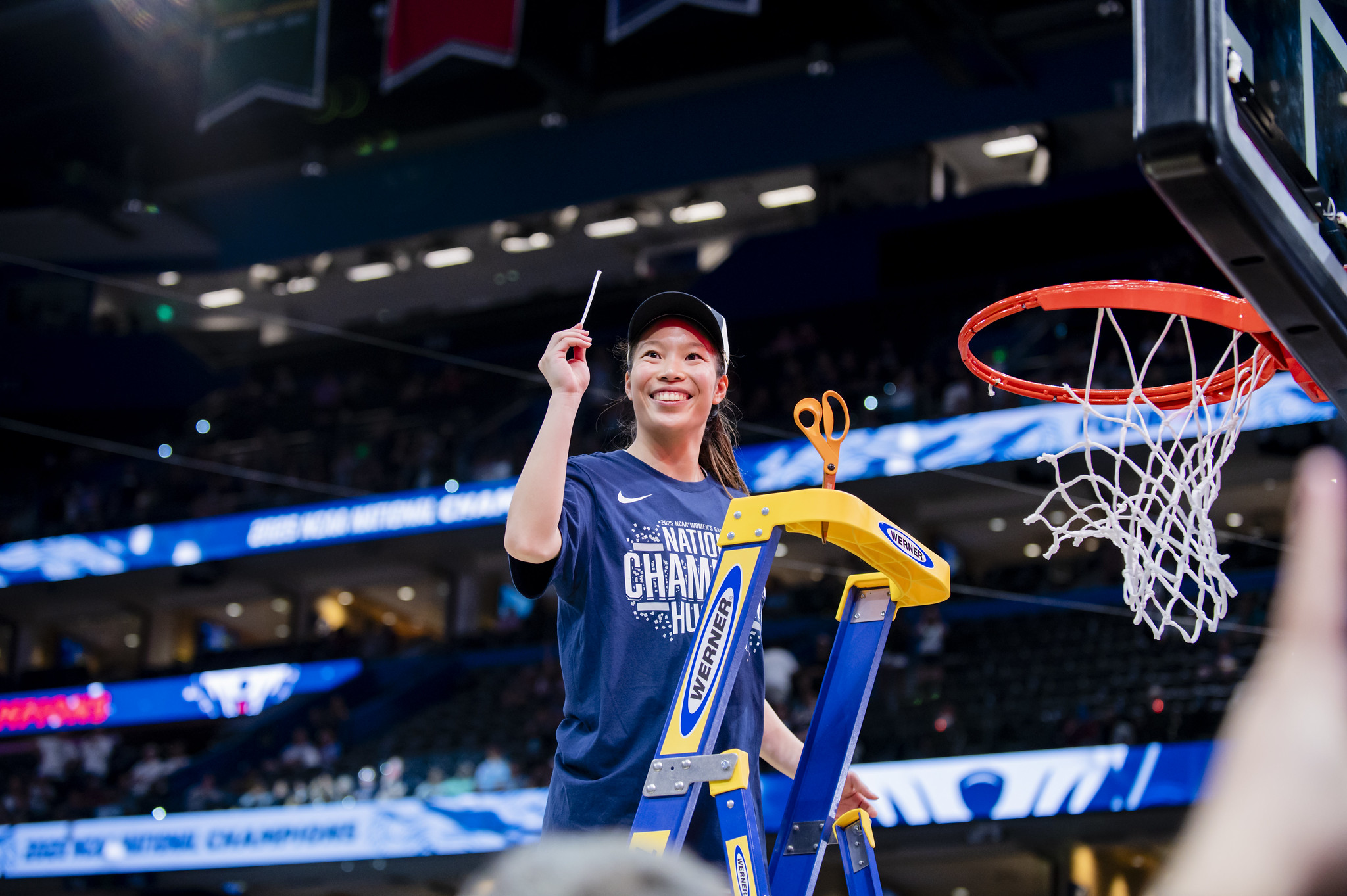 UConn guard Kaitlyn Chen smiles and stands on a ladder. She wears a championship hat and shirt and holds up a piece of the net that she just finished cutting.