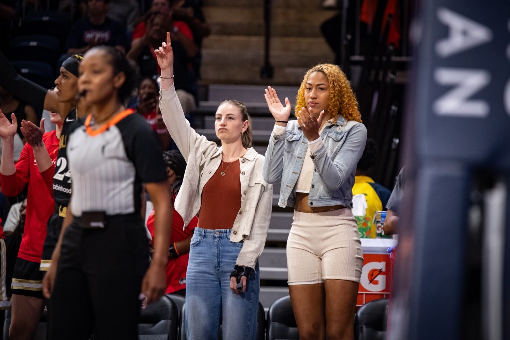Washington Mystics center/forward Shakira Austin stands and claps her hands together on the Mystics' bench. Because she is out with an injury, she is wearing a jean jacket, a cream shirt and cream shorts instead of a uniform.