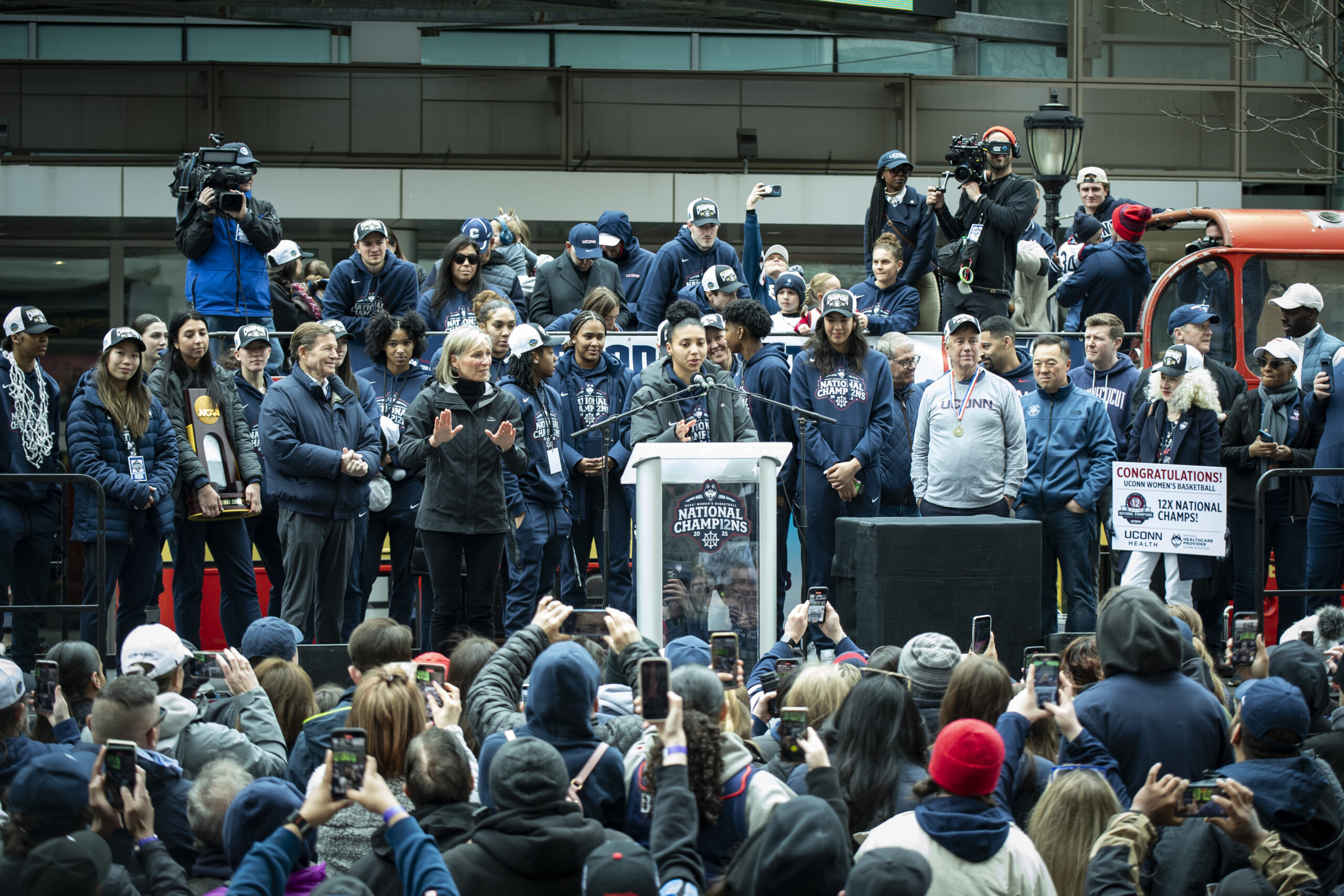 UConn graduate student guard Azzi Fudd stands at the podium with the national champions and UConn logo on it. Her teammates, coaches and prominent Connecticut politicians stand behind her.