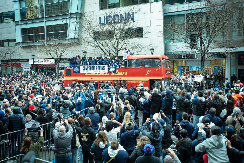UConn takes in the moment from an open-top double-decker bus as fans crowd the streets and capture the moment on their cell phones.